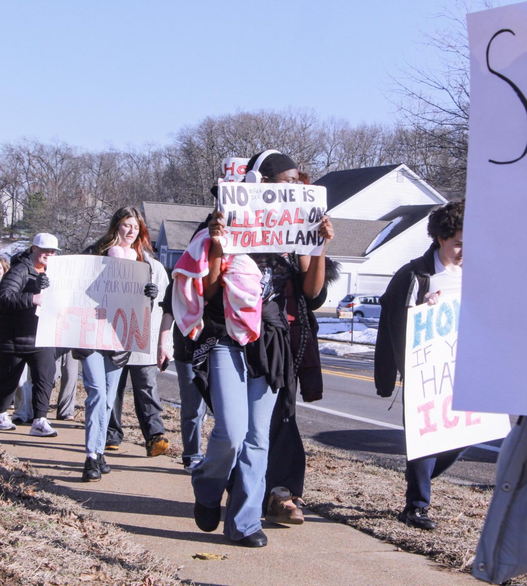 Shielding her face with a sign, sophomore Demayah Balle marches down Clayton Road. Balle's poster reads "No one is illegal on stolen land."